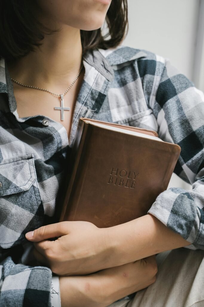 A young woman in a plaid shirt holds a Holy Bible close with a cross necklace visible.